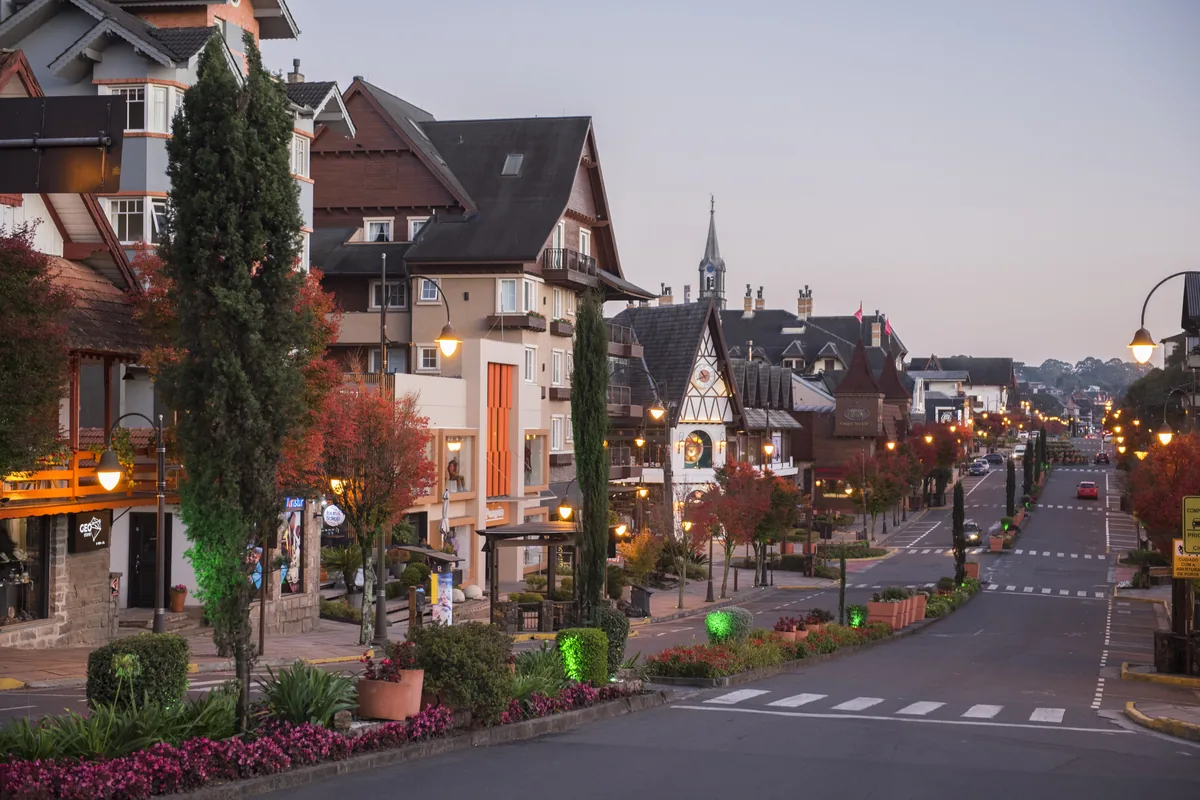 Rua central de Gramado ao entardecer, com casario de arquitetura enxaimel, ciprestes e luminárias acesas