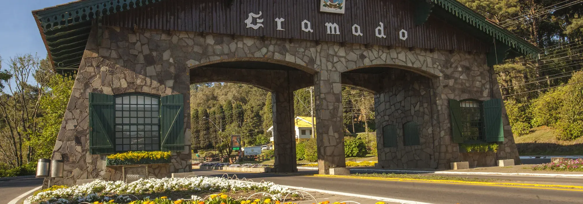 Pórtico de pedra da entrada de Gramado, com telhado verde e canteiro florido em primeiro plano