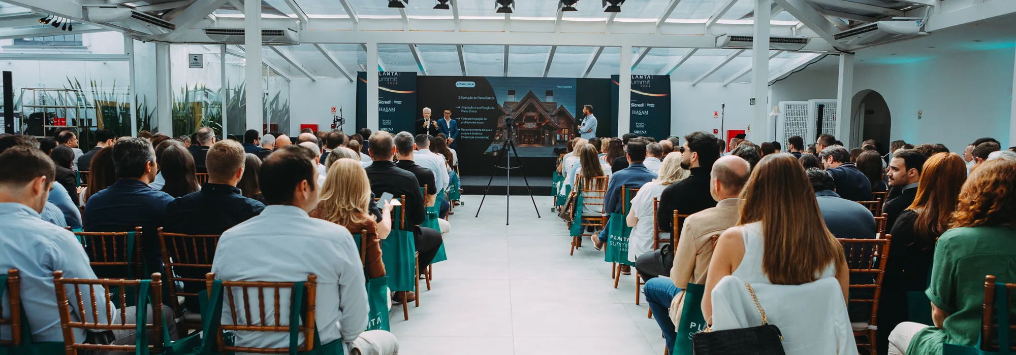 Plateia sentada de frente para o palco durante painel do PLANTA Summit 2026, em salão com estrutura de vidro