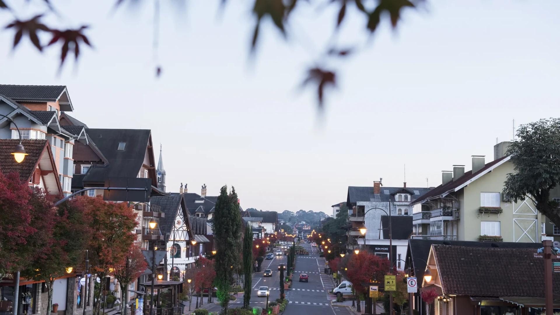 Avenida central de Gramado ao amanhecer, vista entre folhas de acer, com casario de telhados inclinados e luminárias ainda acesas