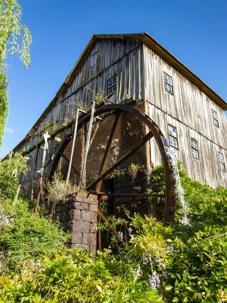 Moinho colonial de madeira com roda d'água em funcionamento, cercado de vegetação, na Serra Gaúcha